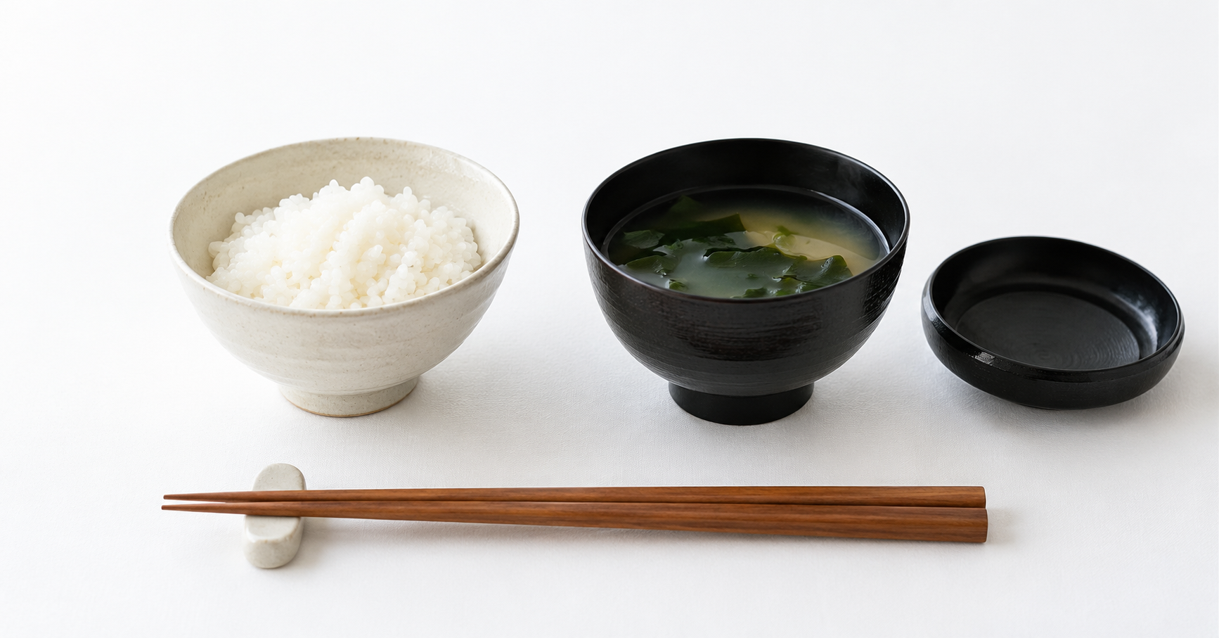 白いご飯茶碗と黒い汁椀、内側を上にして脇へ置いた蓋と箸置きの上の箸
